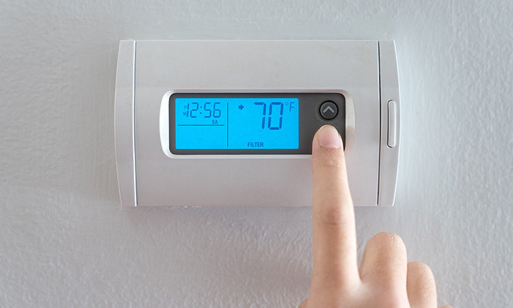 A person is pressing the down button of a wall attached house thermostat with digital display showing temperature 70 degree Fahrenheit for heating, cooling, electricity and gas saving